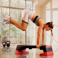 Eine sportliche Frau in weißer Shorts und einem hellblauen Sport-BH macht eine anspruchsvolle Übung auf einem schwarzen und roten Reebok Stepbrett, bei der sie in einer Handstand-ähnlichen Position ihre Knie anzieht. Der helle Trainingsraum hat eine Glasfront mit Blick ins Grüne.