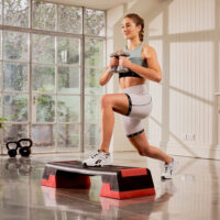 Eine Frau in Sportkleidung führt einen Step-Übung mit Kurzhanteln auf einem schwarzen und roten Reebok Stepbrett aus. Sie hat ein Lächeln im Gesicht und trainiert in einem Raum mit großer Fensterfront und weißen Holzwänden.